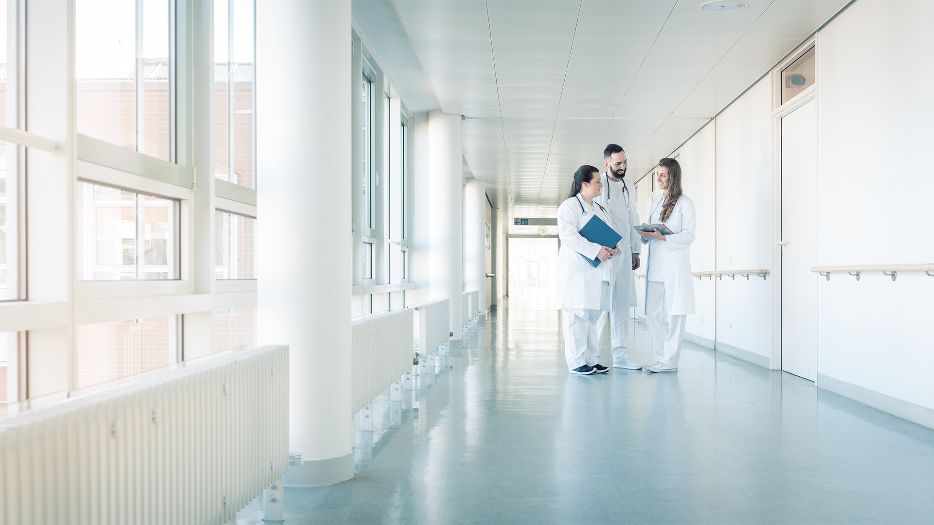 Ein Team aus zwei Ärztinnen und einem Arzt stehen in weißer Bekleidung auf einem leeren Krankenhausflur und unterhalten sich
